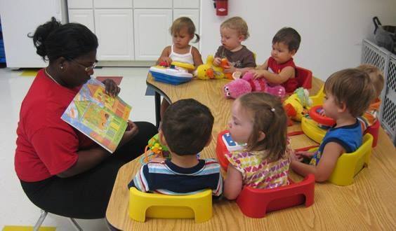 Teacher reads a book to children seated at a table with toys in a classroom.