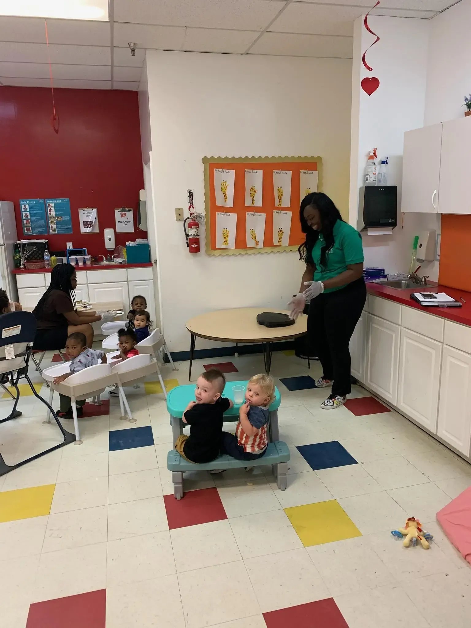 Childcare room: Caregiver tending table, children seated nearby. Red and white cabinets, colorful floor tiles.