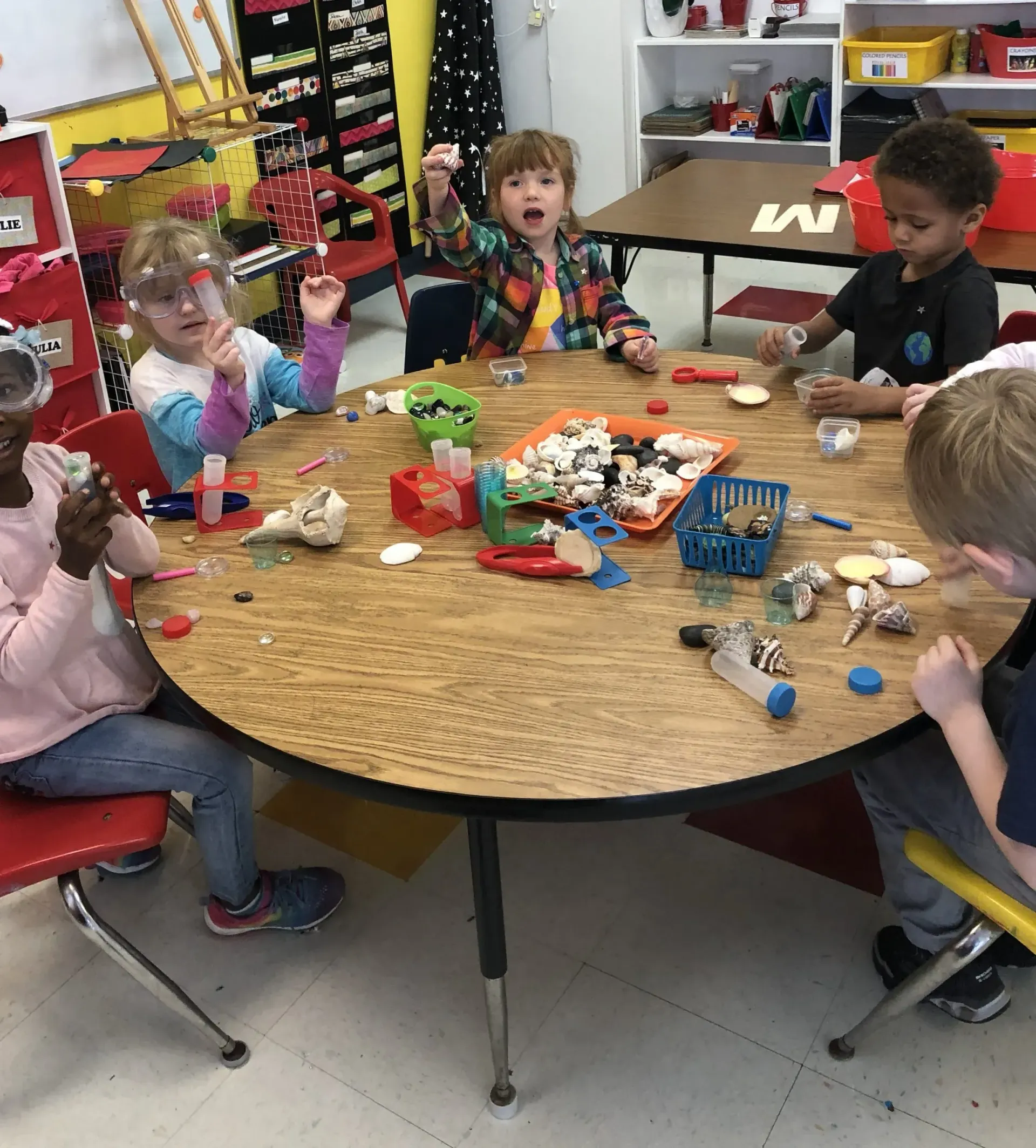 Children seated around a table, exploring various objects. Classroom setting with colorful toys and activities.
