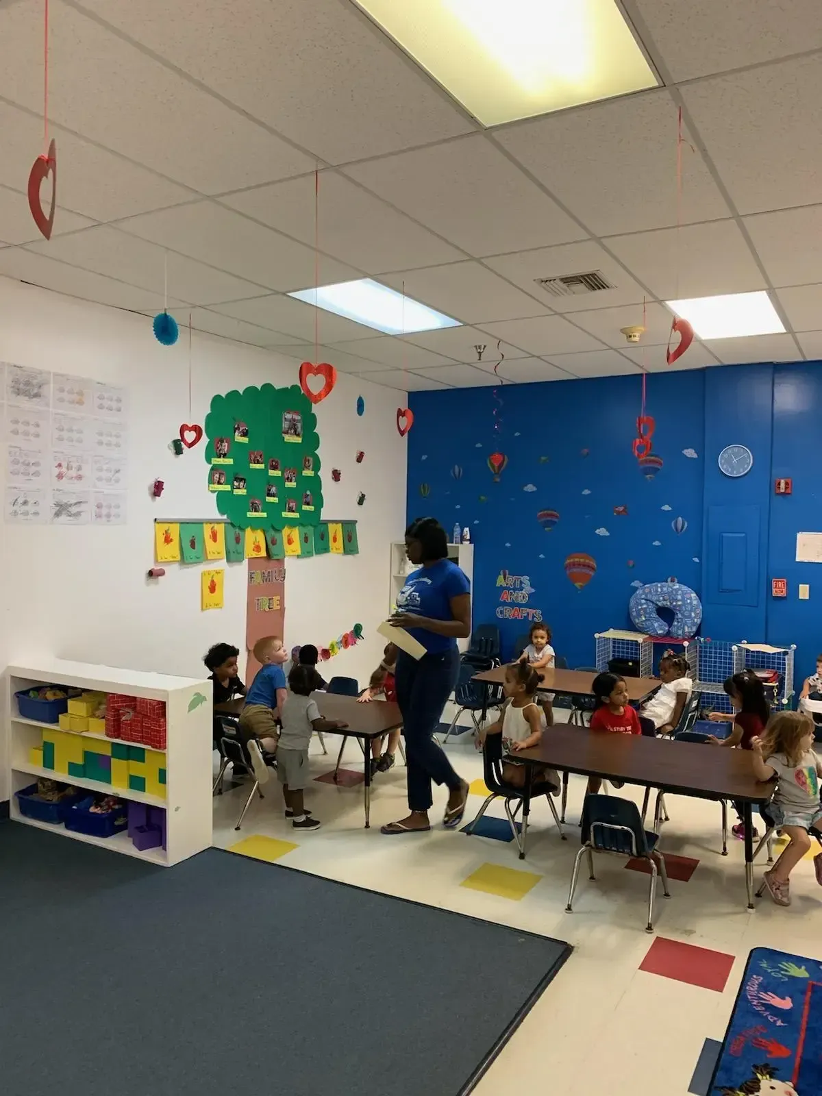 Teacher in classroom with children at desks, colorful decorations on walls.