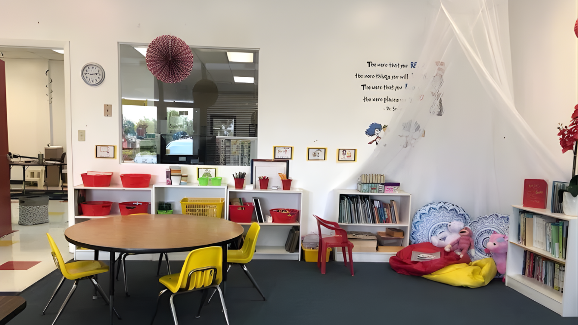 A colorful classroom with tables, shelves, and reading nook. Red, yellow, and white predominate.