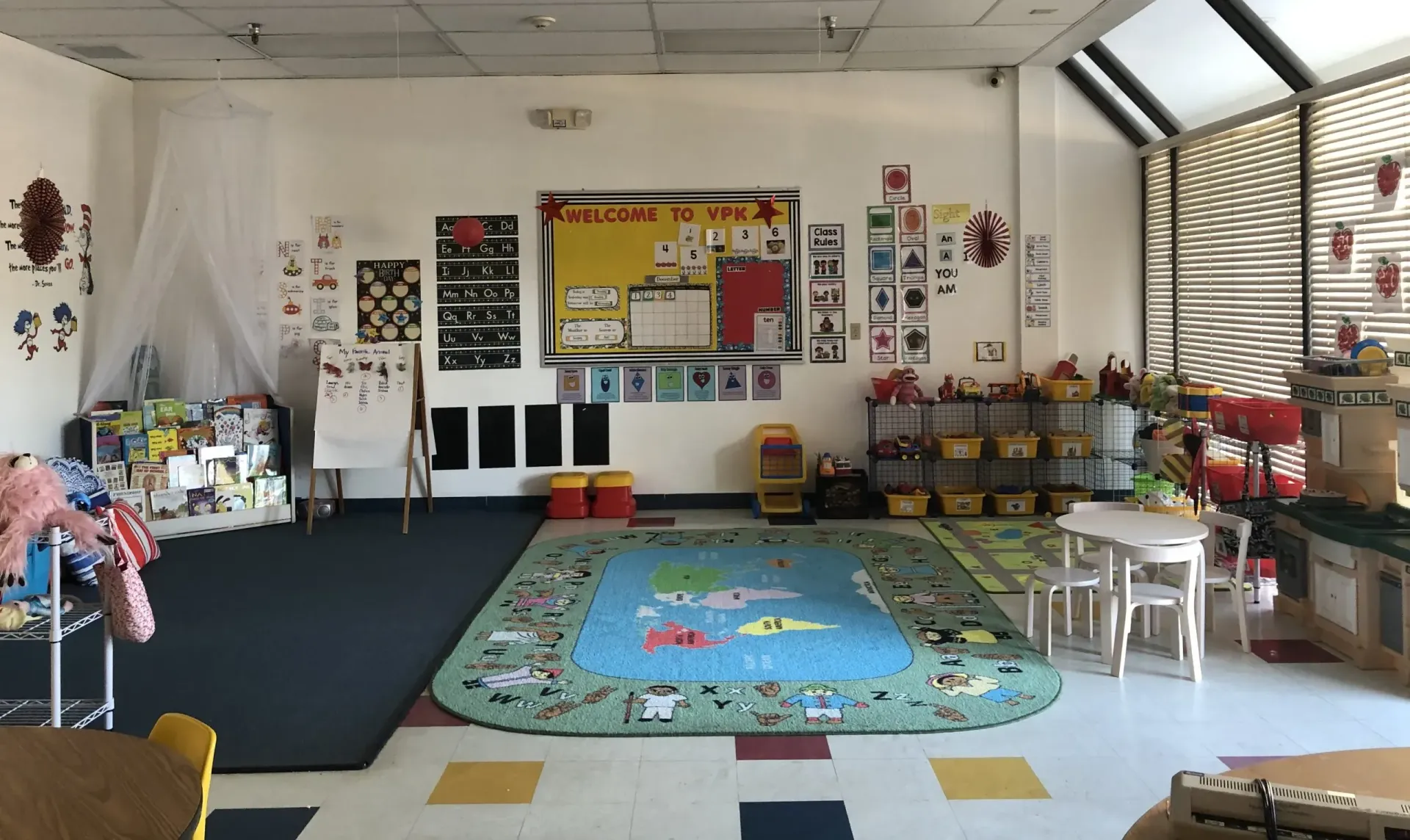 Classroom interior with rug, tables, and learning materials. Bulletin boards and windows are visible.