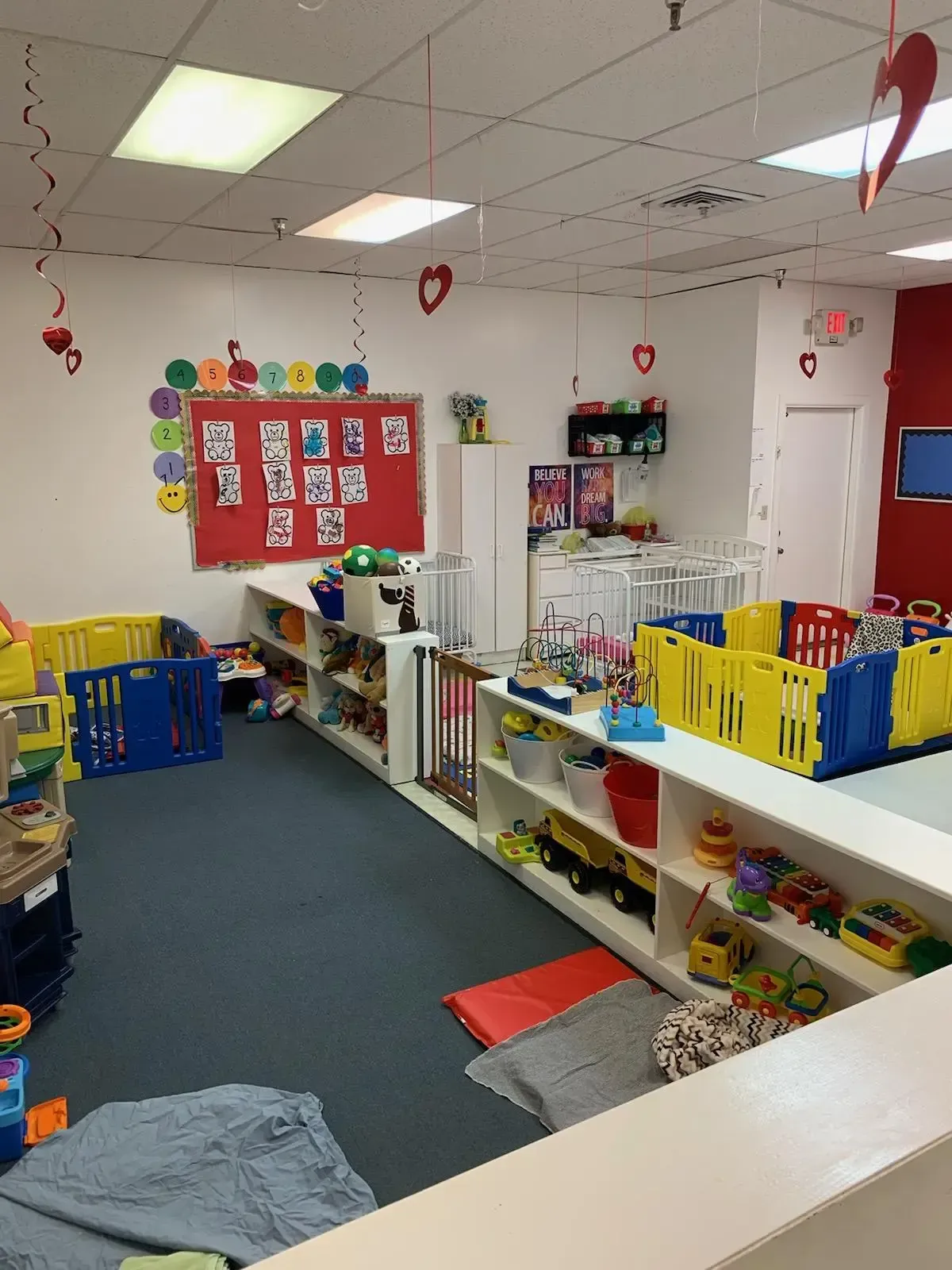 Daycare room with toys, cribs, and educational wall displays. White, red, and blue colors dominate the scene.