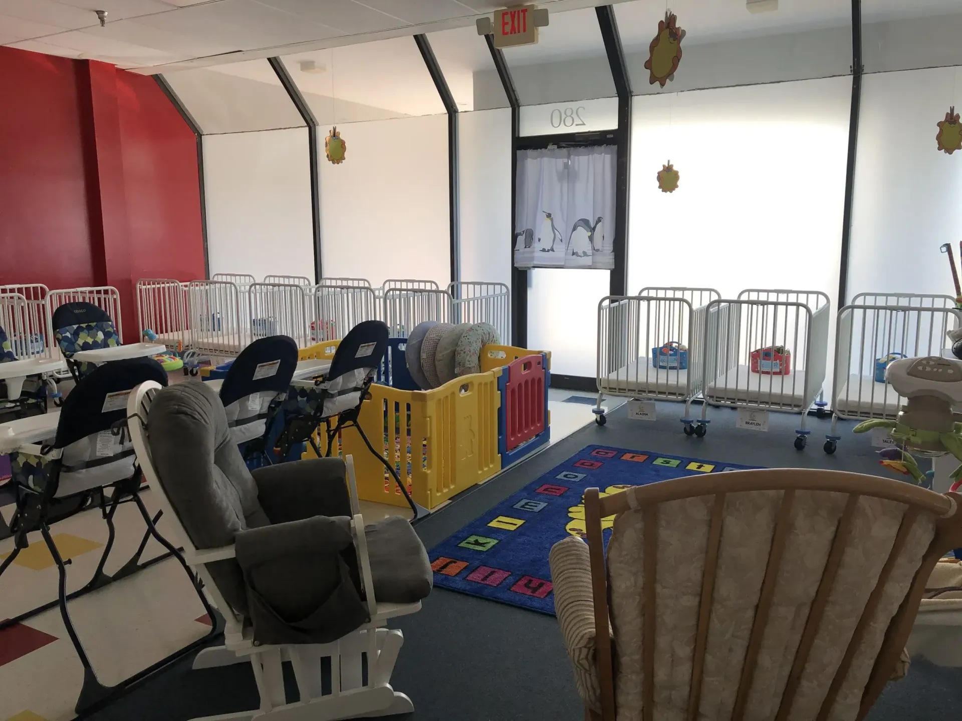 Nursery room with cribs, play area, and chairs. Red and white walls, door to the right.