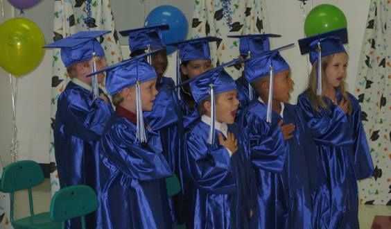 Children in blue graduation gowns and caps, singing, with balloons in background.