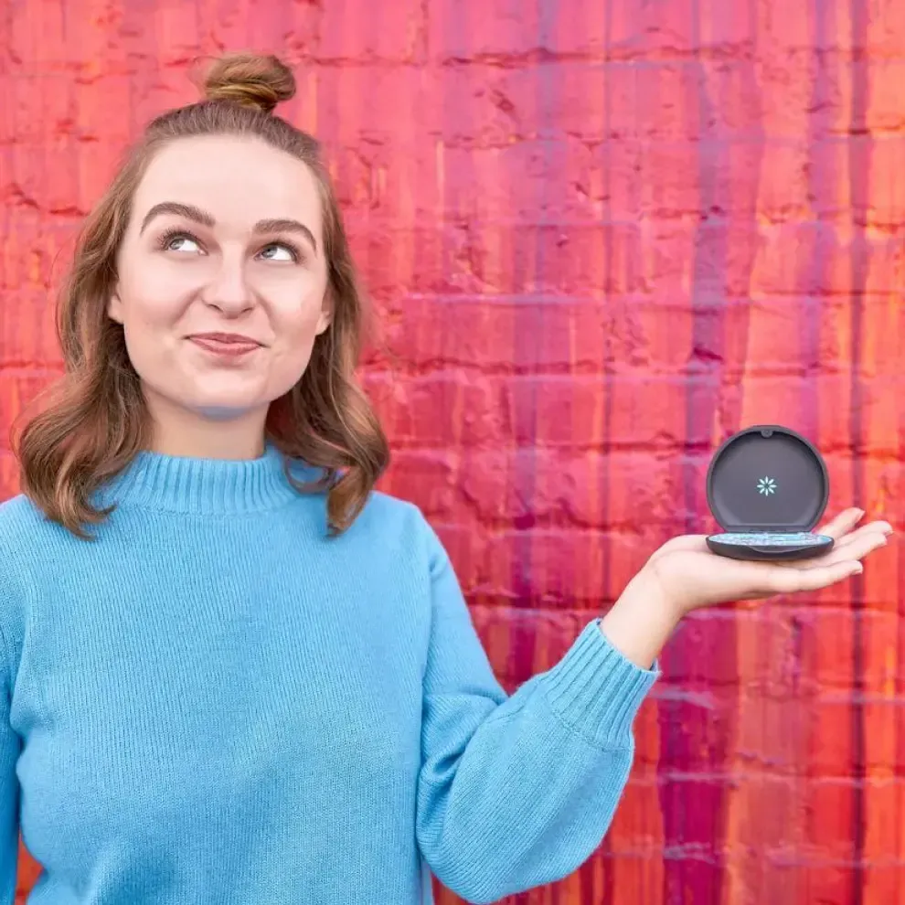 A woman in a blue sweater is holding a round object in her hand in front of a red brick wall.