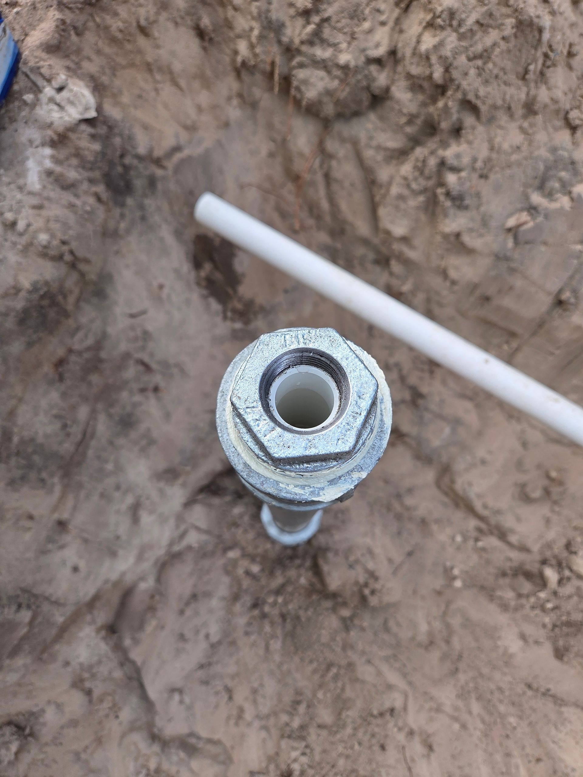 Metal well cap and pipe in a sandy trench with a length of white PVC pipe nearby.