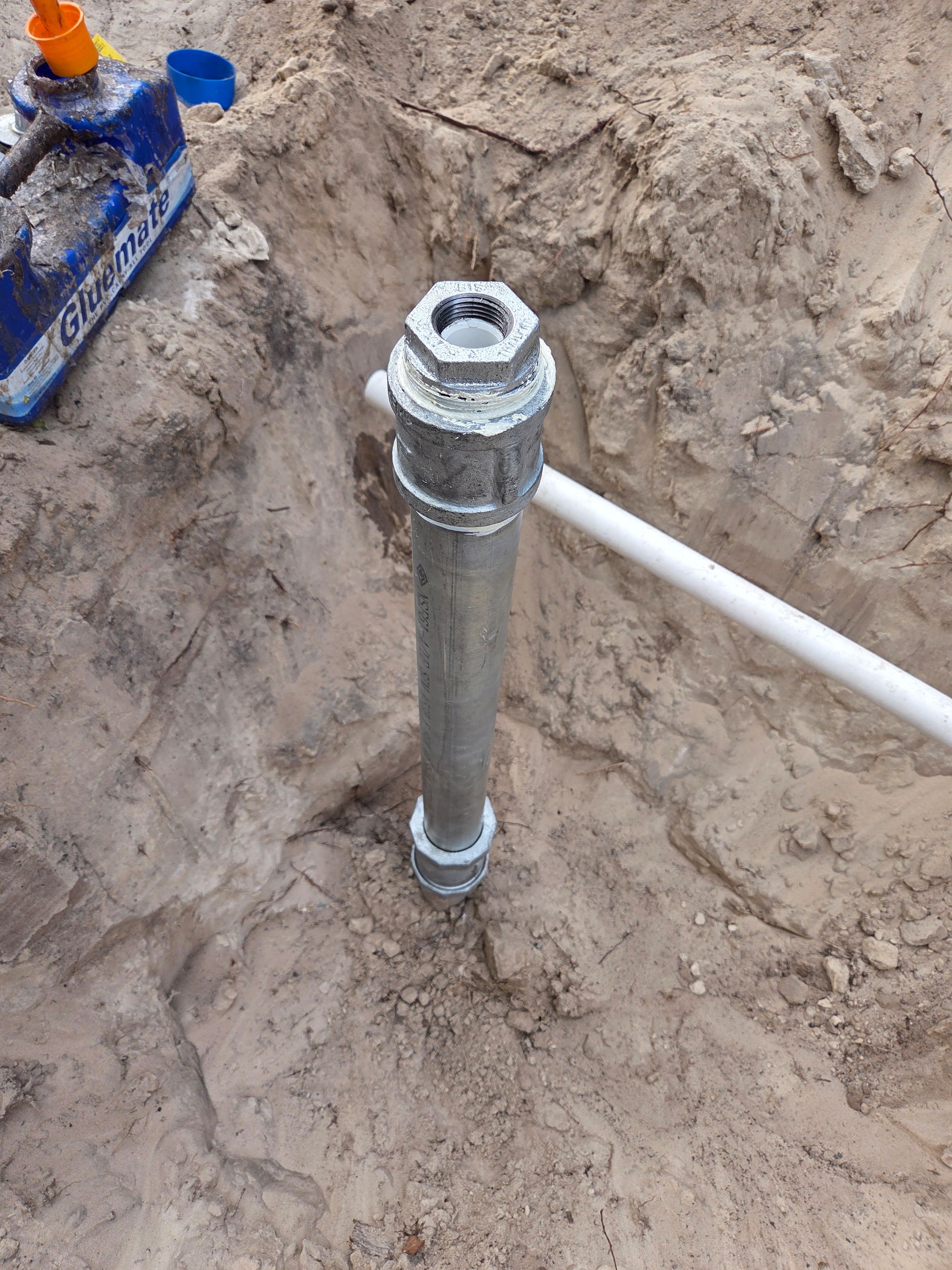 Metal well pipe installed in sandy soil with a white PVC pipe extending from it, and tools nearby.