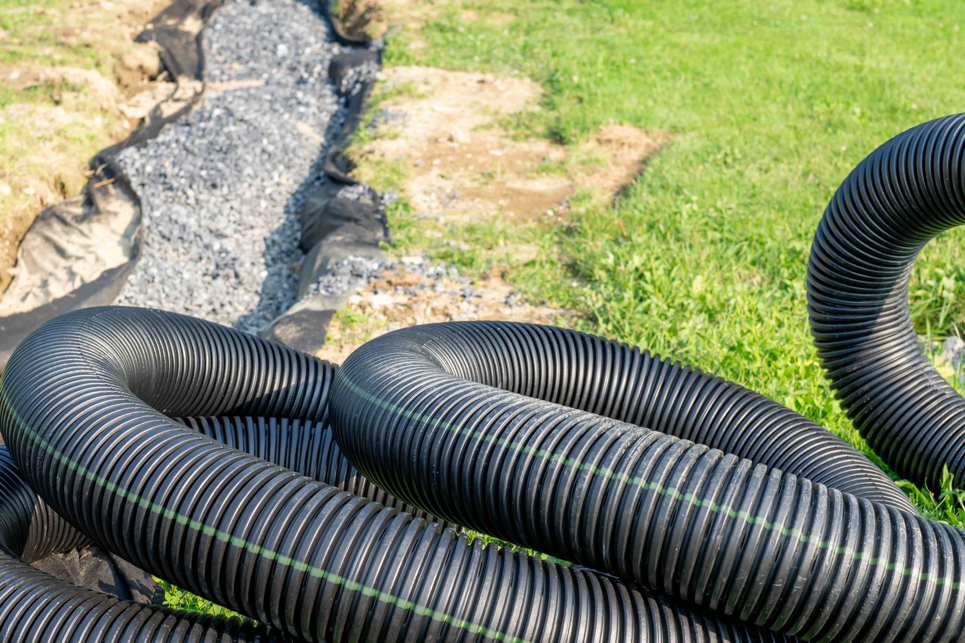 Black corrugated drainage pipe coiled near a trench filled with gravel and fabric, on green grass.