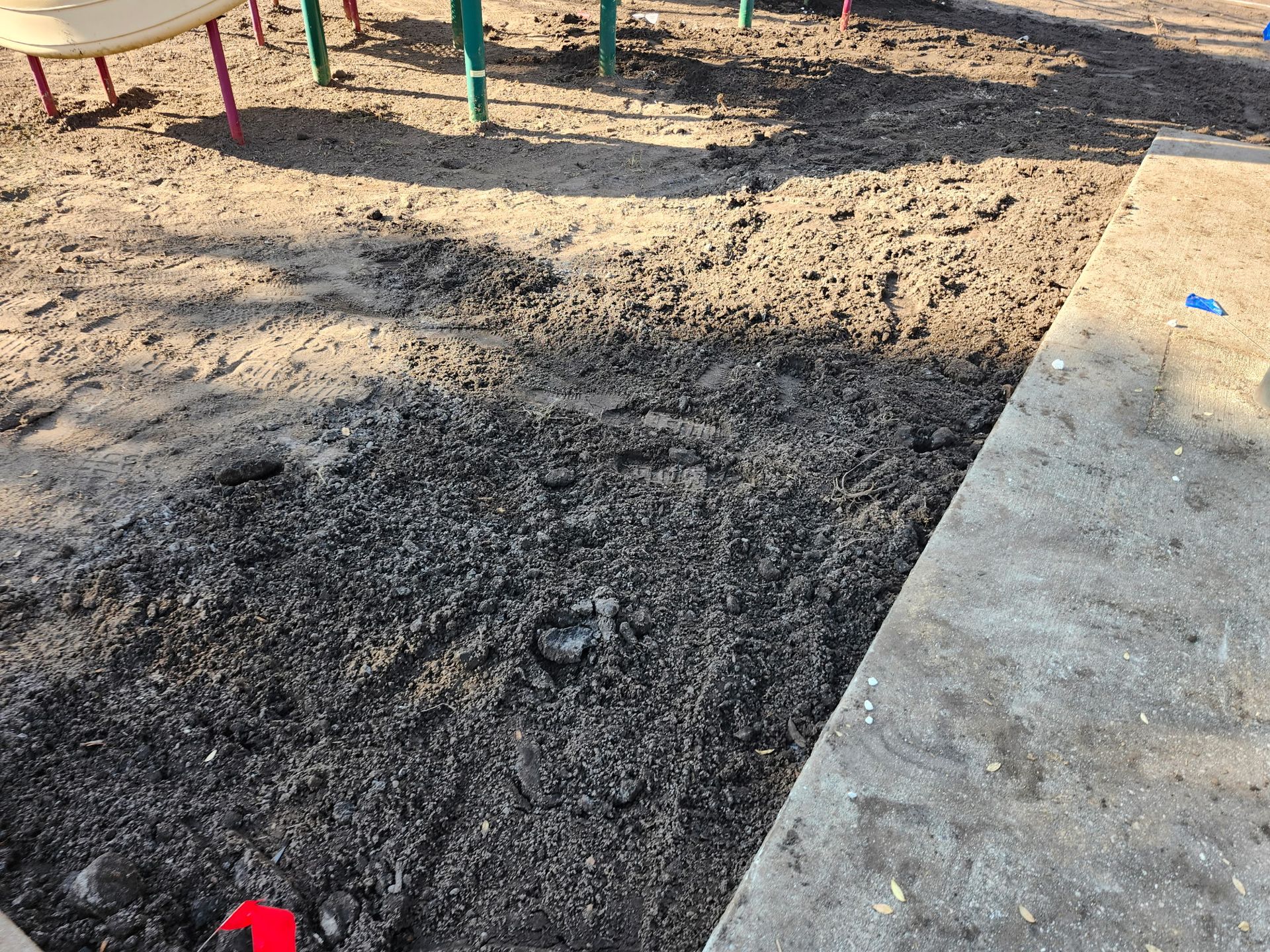Dark, loose material, possibly mulch, next to concrete border and playground equipment.