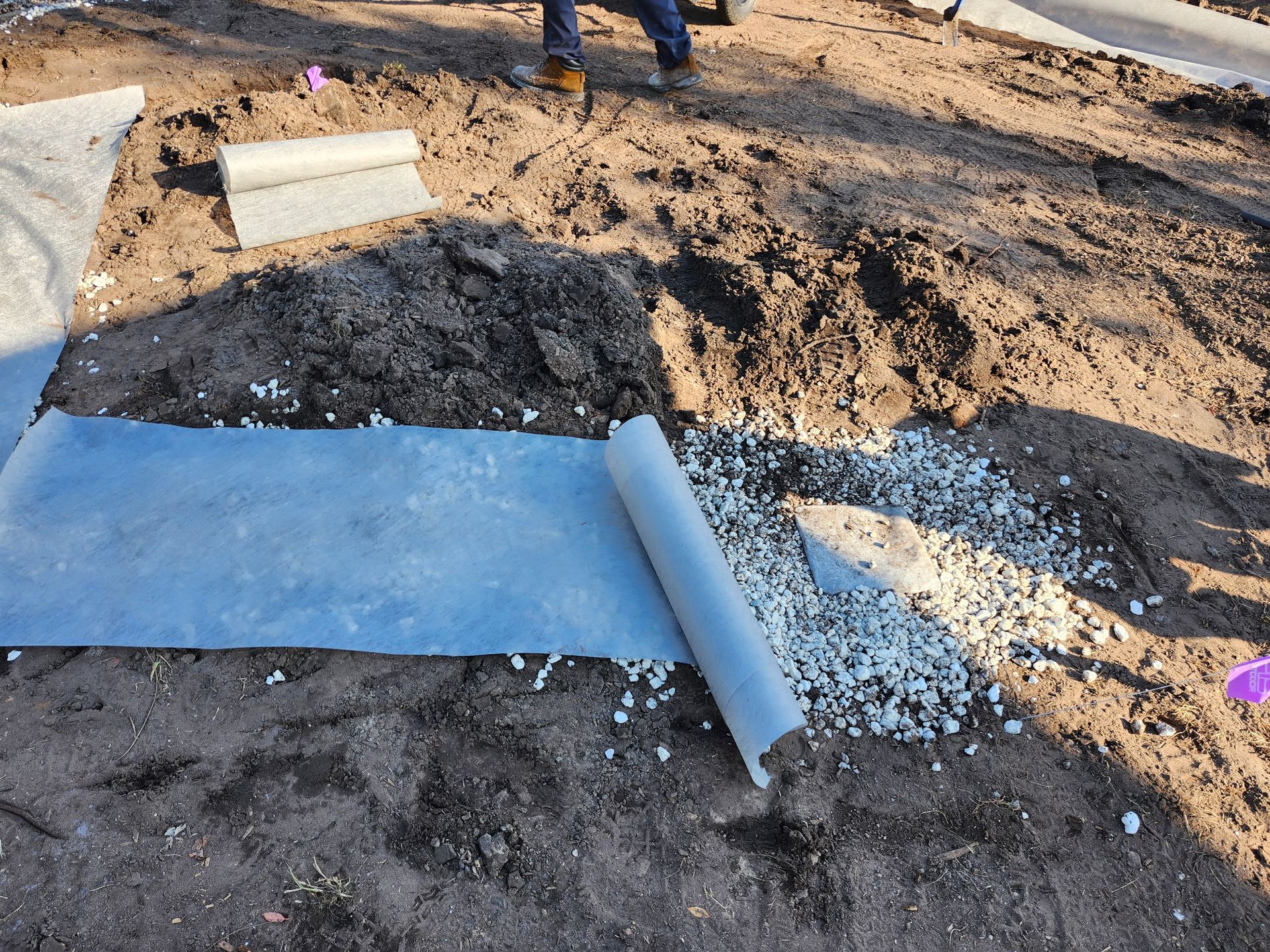 Roll of landscape fabric being unrolled on a patch of soil with gravel. Person standing nearby.