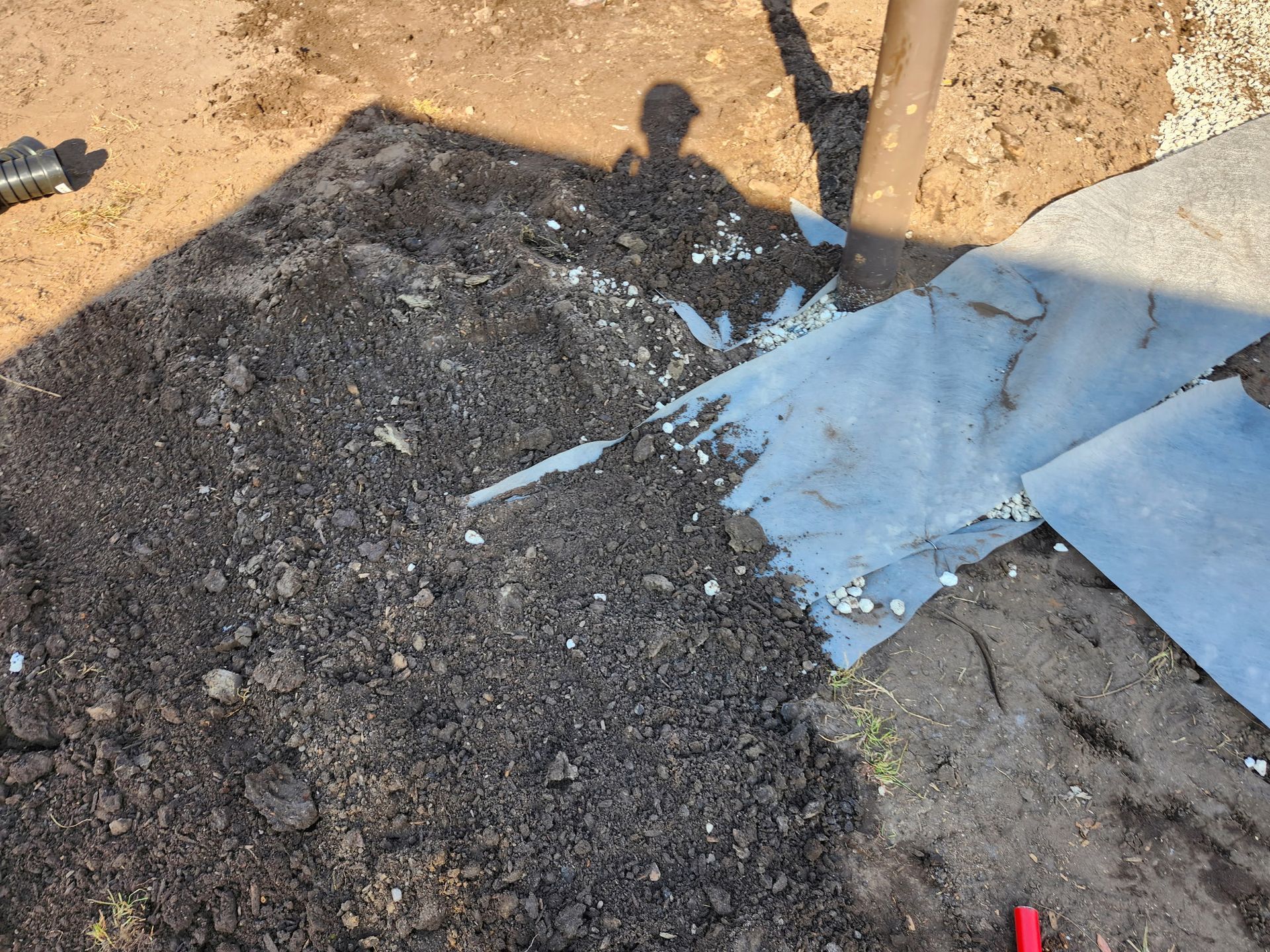 Pile of dark soil, torn blue plastic, and small rocks on a dirt surface. Person's shadow visible.