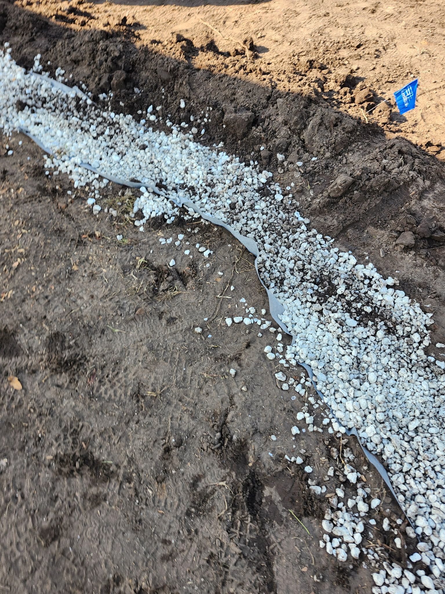 Long trench filled with small, light-colored stones, bordered by dark soil and a blue marker.