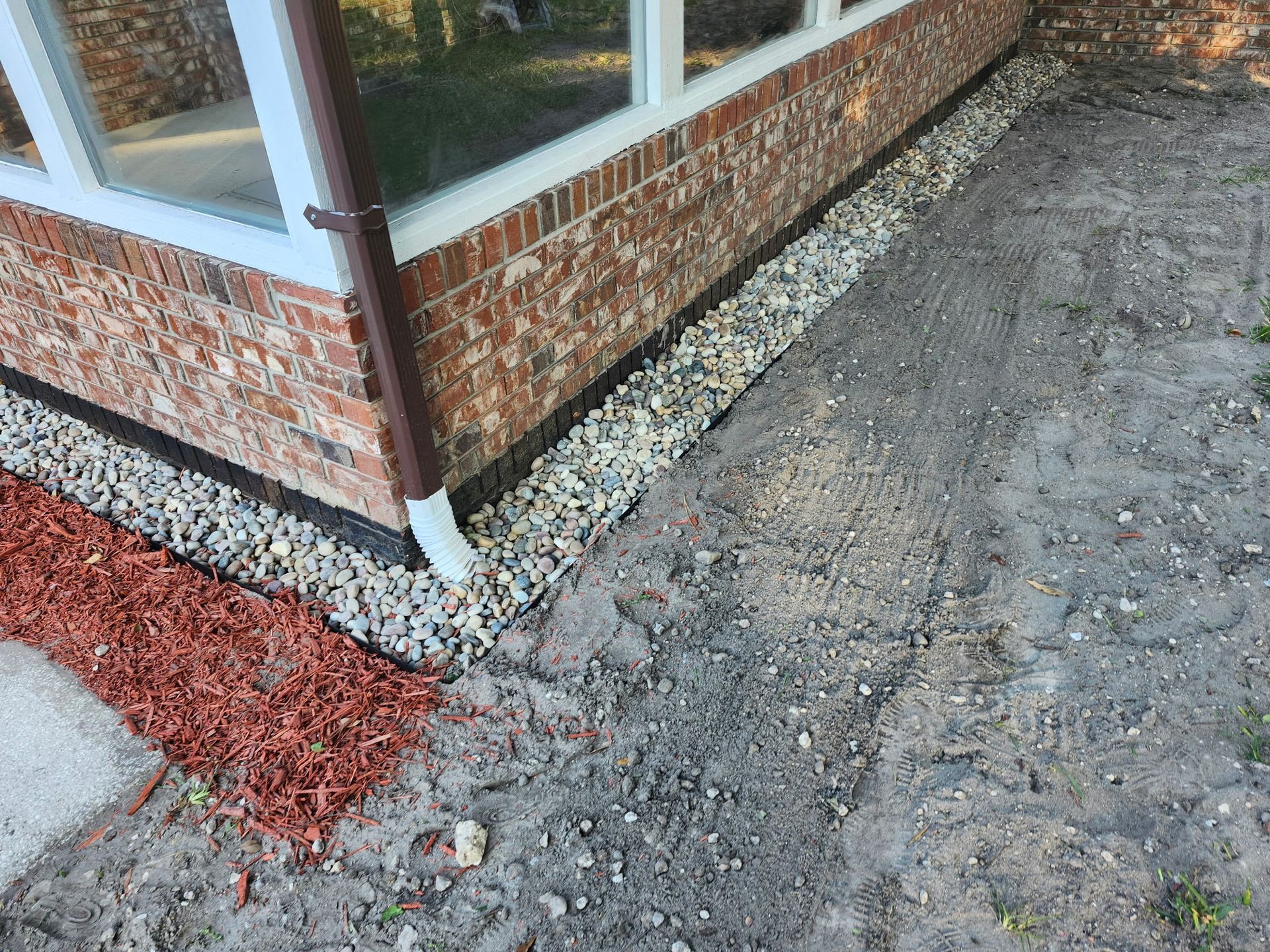 Brick building with gravel and mulch along the foundation. Brown downspout and dirt area.