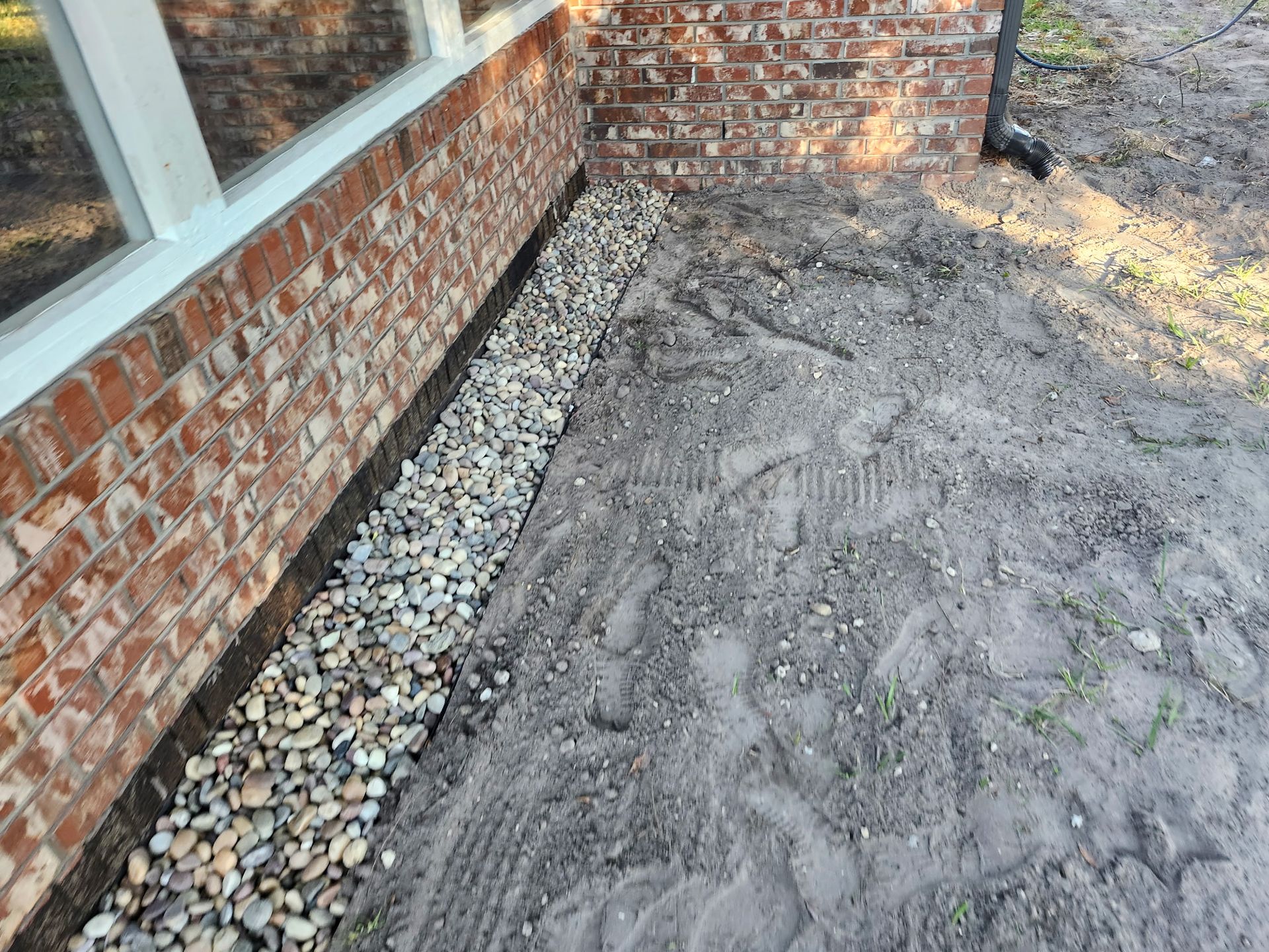 A gravel-filled trench next to a brick building foundation, beside a window and bare dirt.
