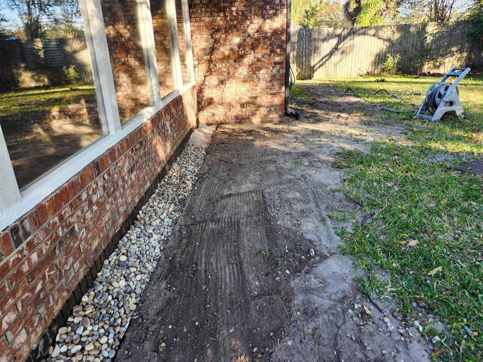 Exterior view: brick wall, dirt path with gravel border next to a window; grass and backyard in the background.