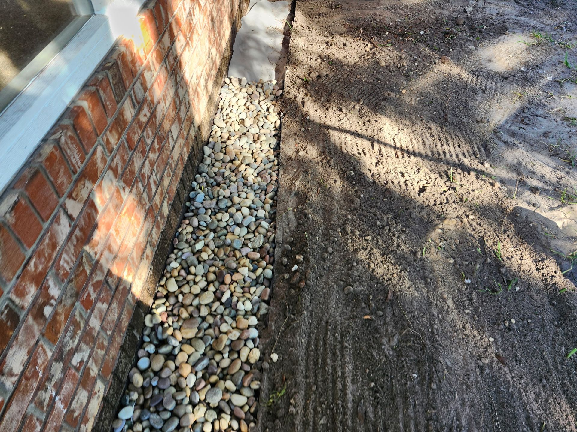 Gravel and dirt border a brick wall and a paved section. Sunny outdoor setting.