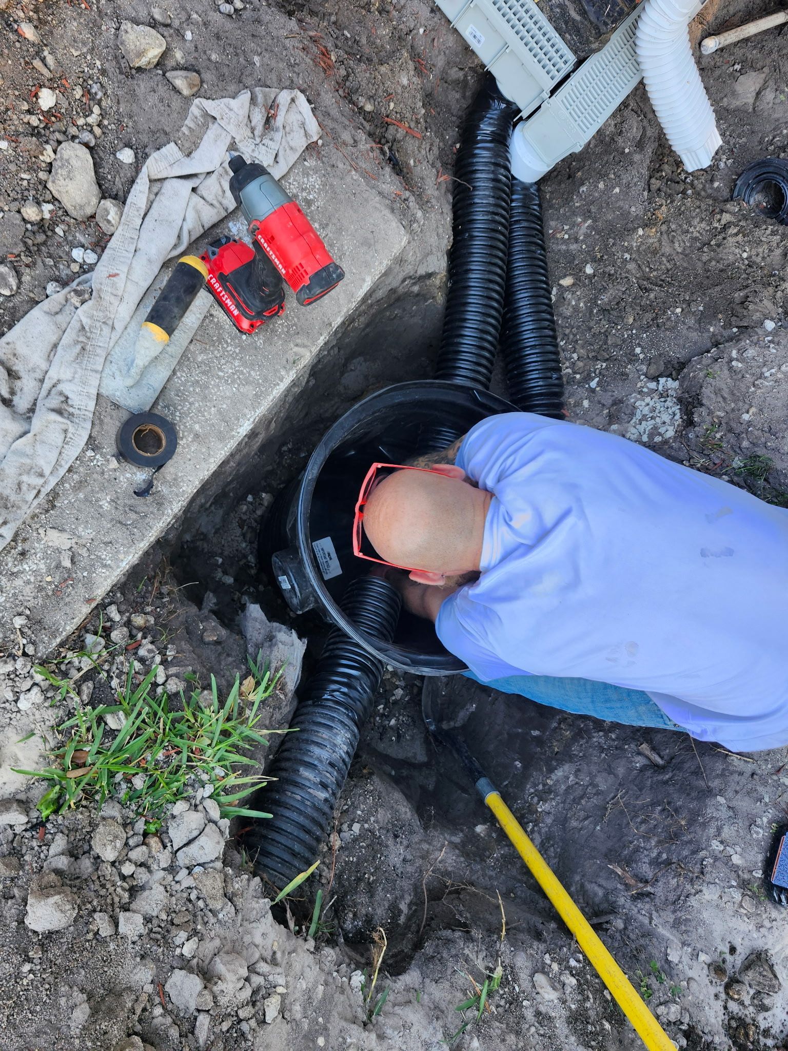 Person in light-blue shirt working on a drainage system in a hole in the ground, with black pipes and tools visible.