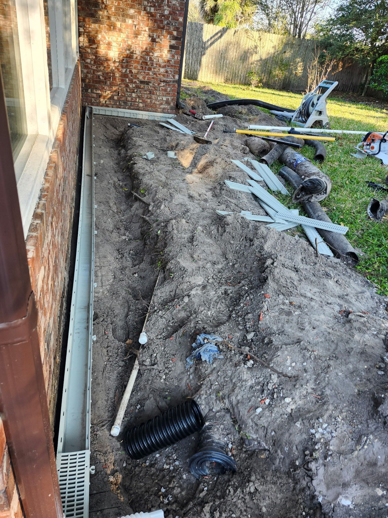 Trench dug along a brick wall for drainage, with pipes and construction materials visible. Outdoors.