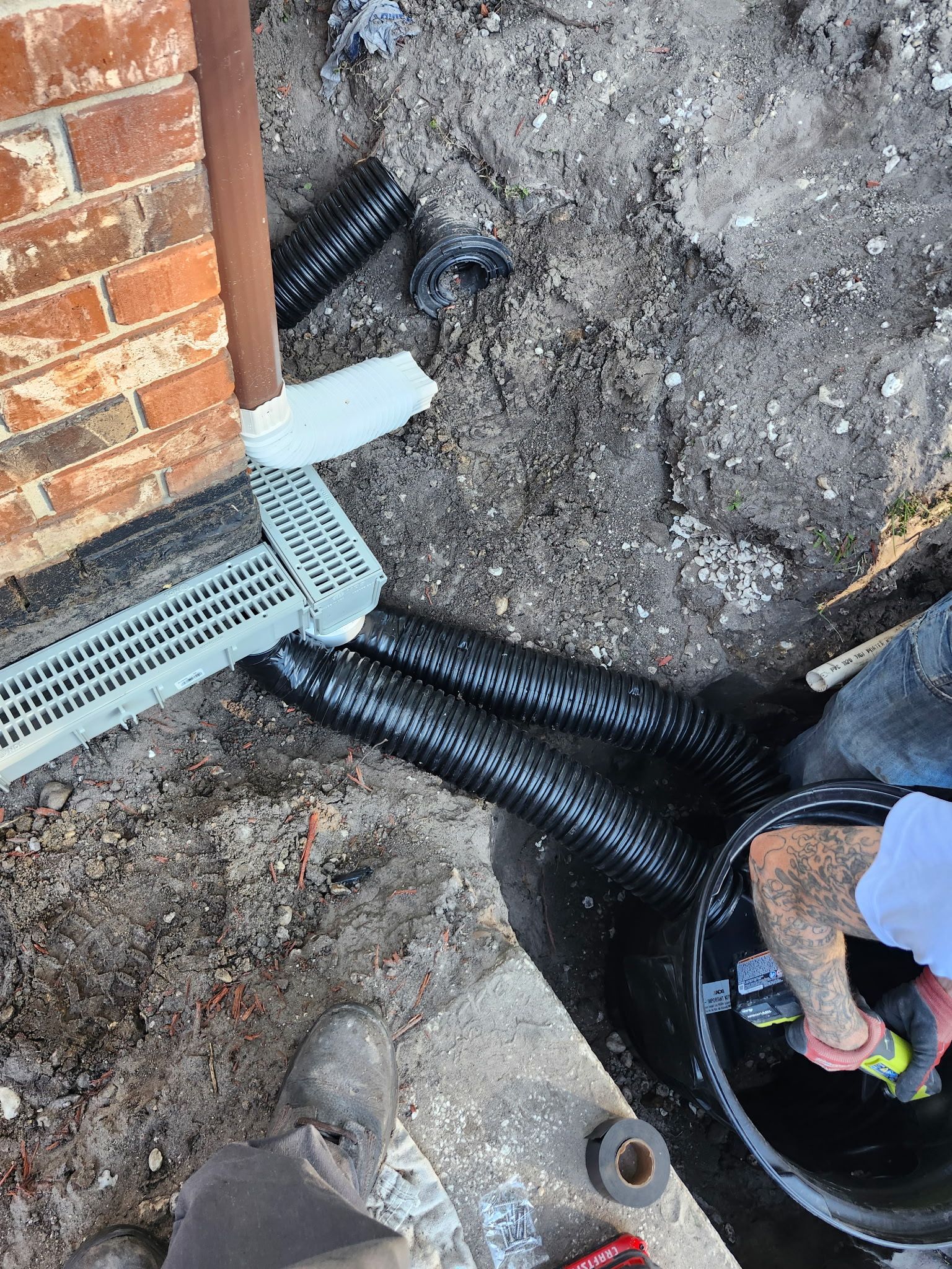 Drainage system installation by a brick wall; black corrugated pipes connect to a grate. Person working in a hole.