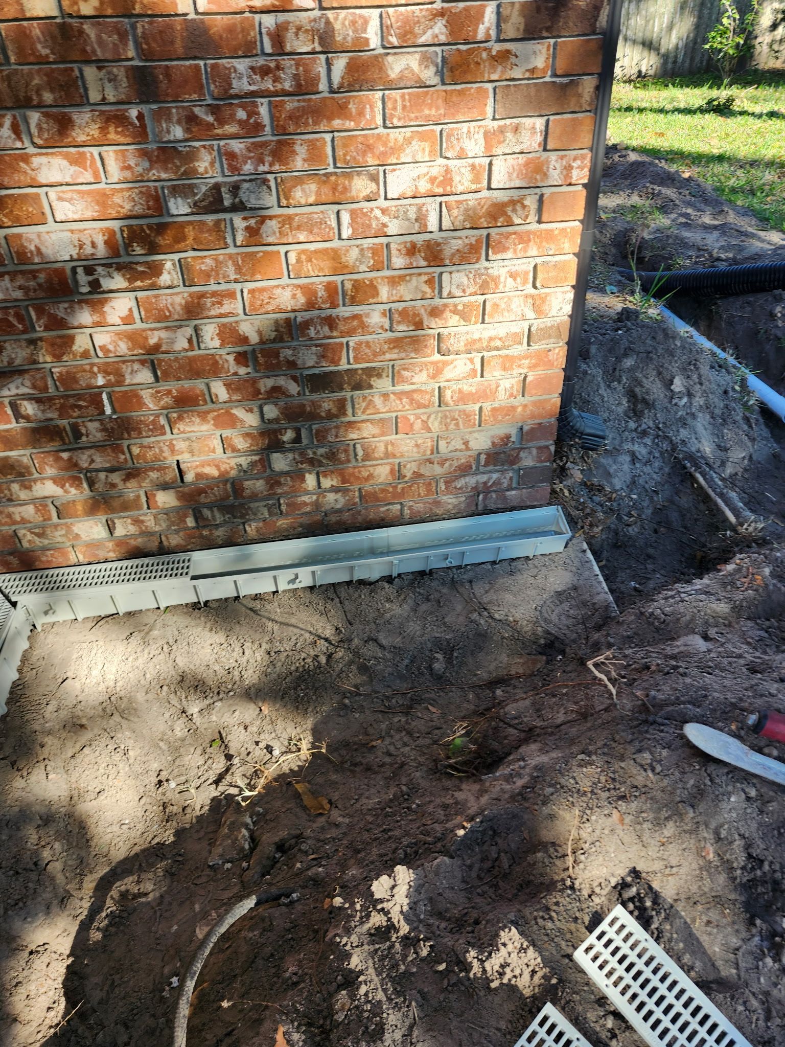 Brick wall next to a trench with a drainage channel installed. Dirt and grass in the surrounding area.