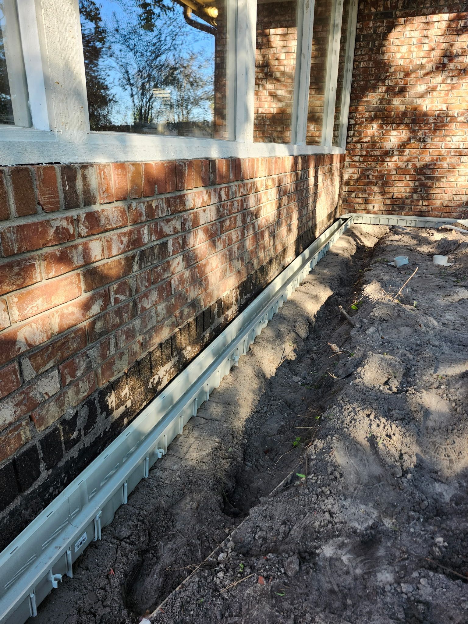 Metal trench drain alongside a brick building, set in dirt.