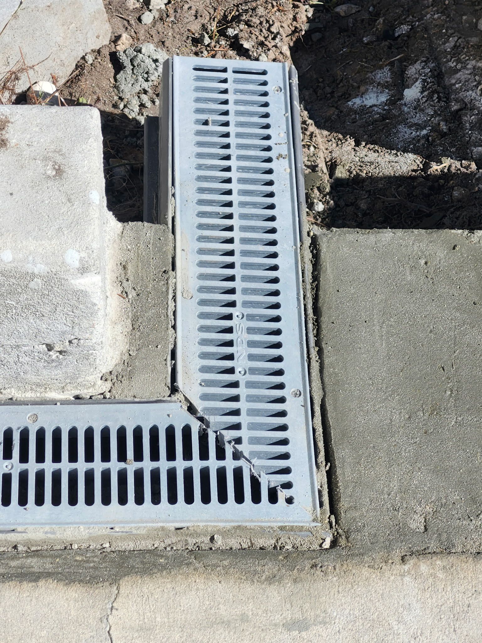 L-shaped metal drain with slotted cover in concrete, at the edge of a sidewalk.