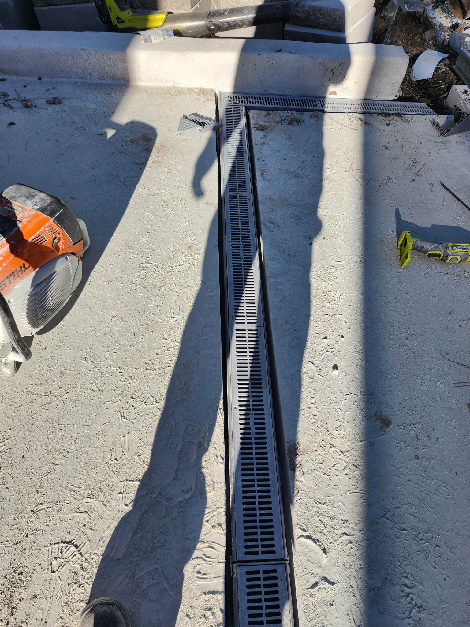 A concrete trench drain with a dark grate in a construction setting. A circular saw and other tools are visible.