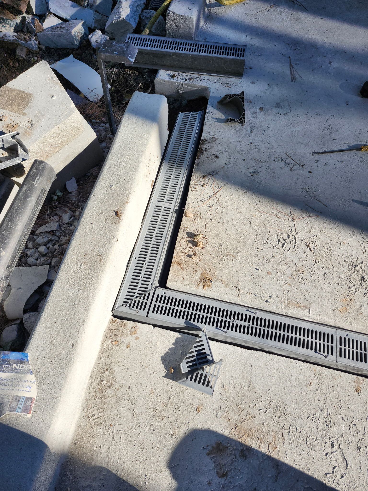 Concrete work with a gray grate drainage system installed at a corner, debris visible.