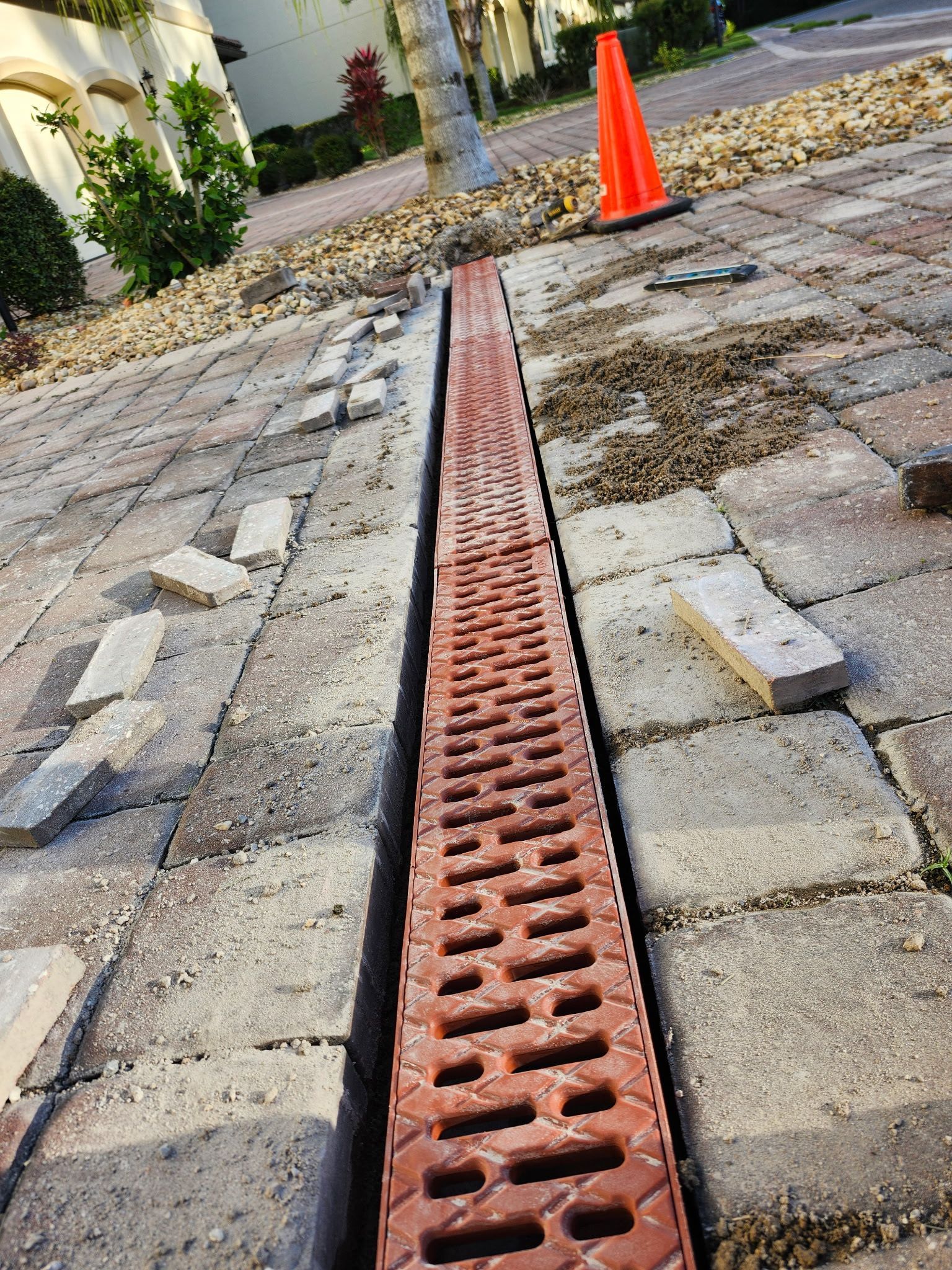A newly installed red brick drainage system in a paved driveway. Construction materials and an orange cone are present.