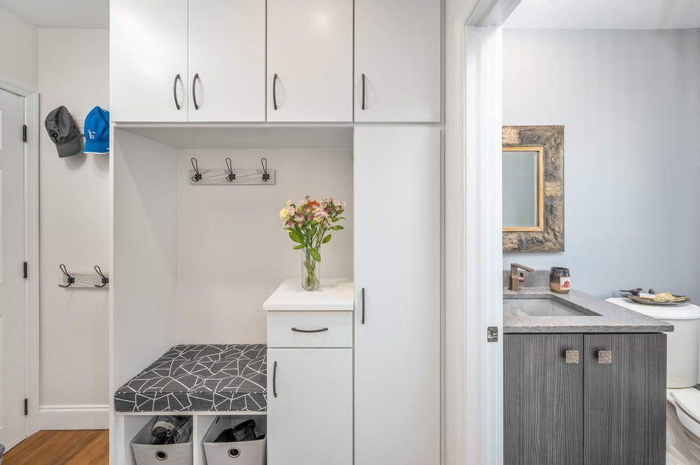 A hallway with white cabinets , a bench , and a mirror.