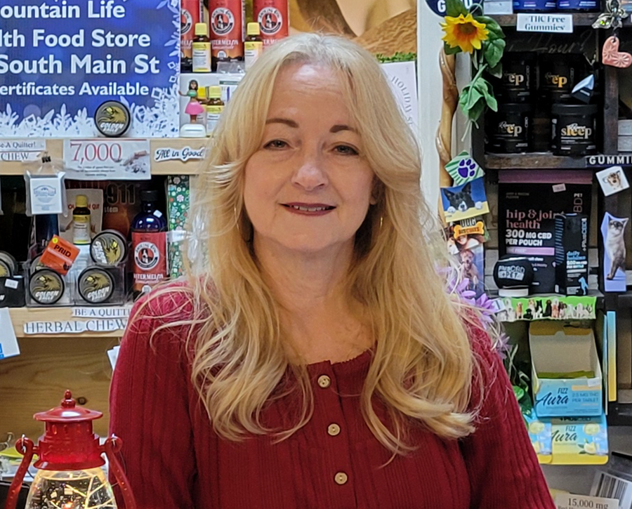 Woman with blonde hair smiles in a health food store, wearing a red sweater. Shelves with products behind her.