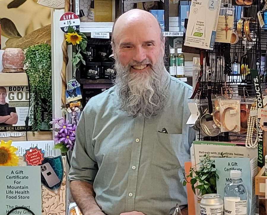 Man with a long, gray beard smiling, standing in a shop filled with products.