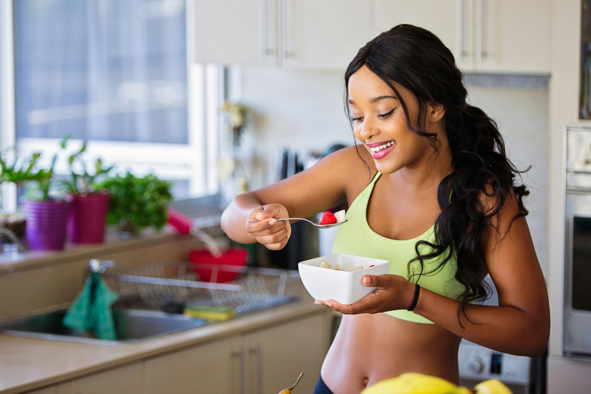 Woman eating fruit in a kitchen; smiling, holding a bowl and fork, wearing a green sports bra.