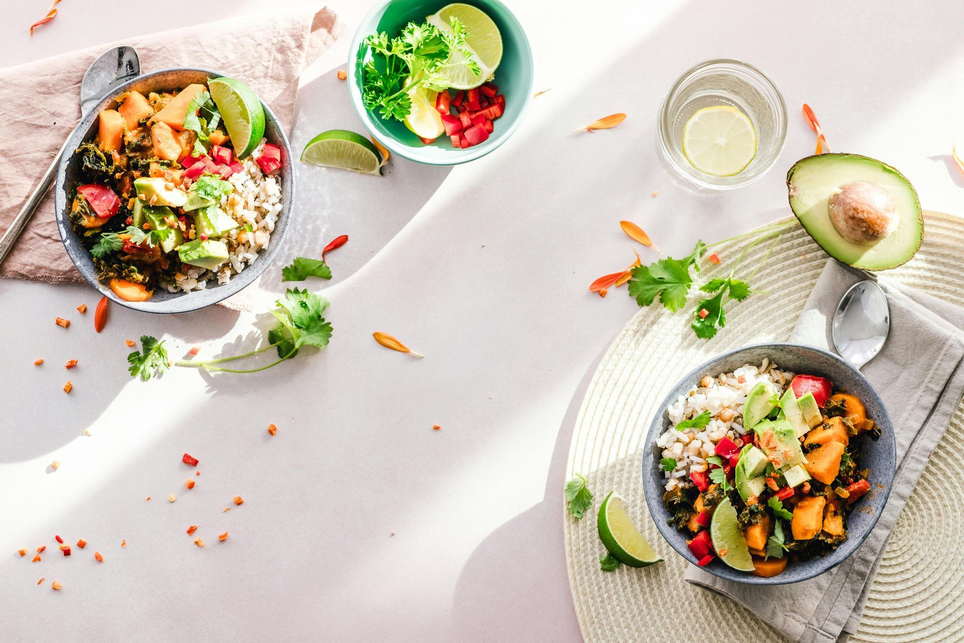 Bowls of colorful food on a white table with lime slices, avocado, and water glass.