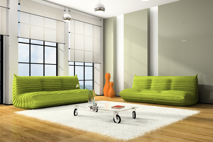 Bright green couches on timber floor, around industrial glass coffee table with wheels on white shaggy rug. Semi see-through blinds pulled half way down at different levels framing black framed windows.