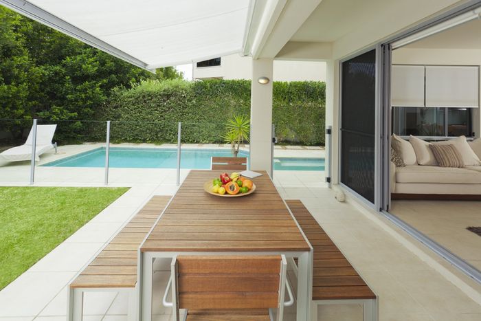 Wood and metal outdoor table and chair set, with full fruit bowl on top, overlooking glass fenced pool surrounded by green hedge and green grass.