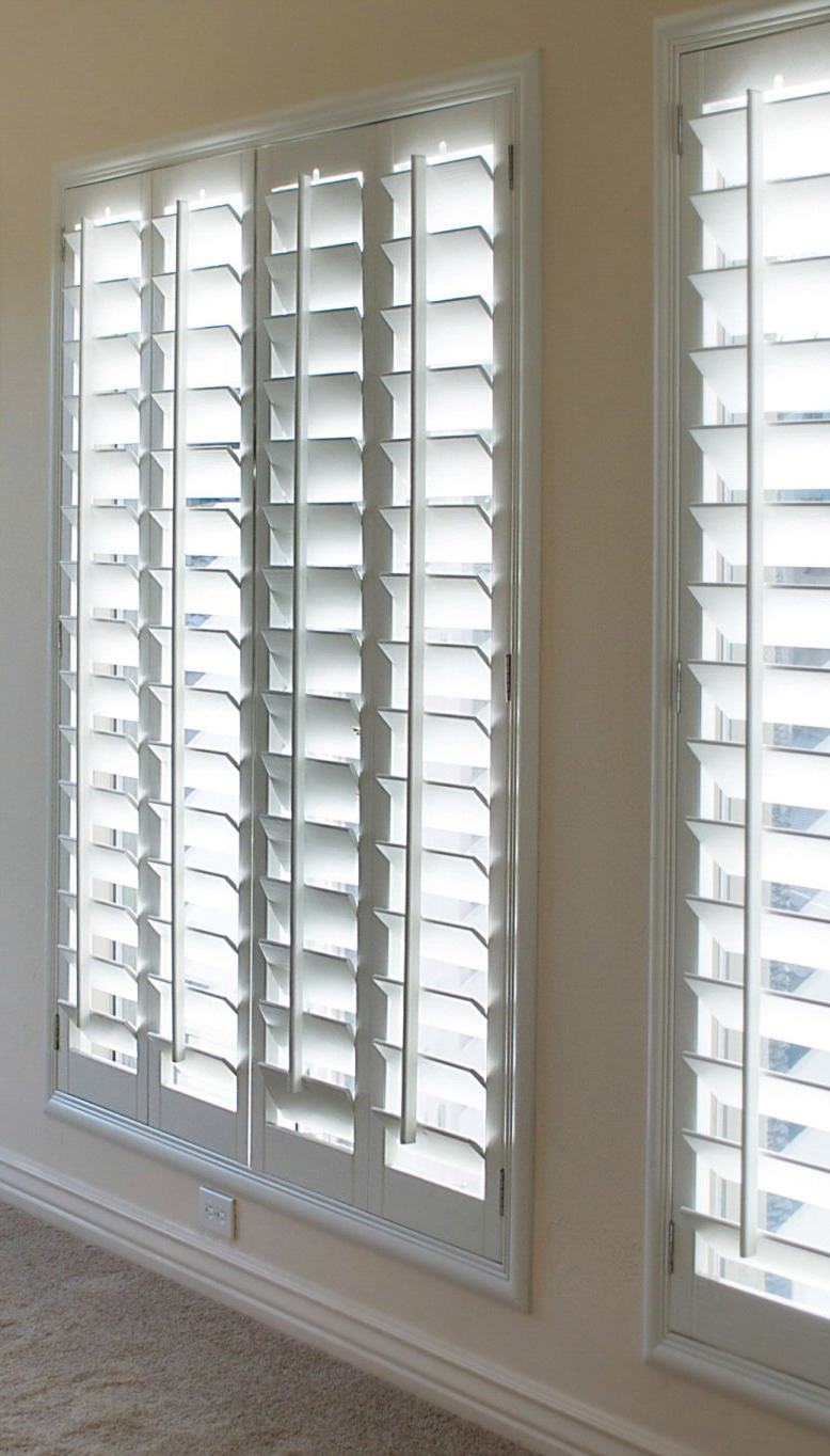 Large set of full length plantation shutters, slightly open to let in sunlight to carpeted room.