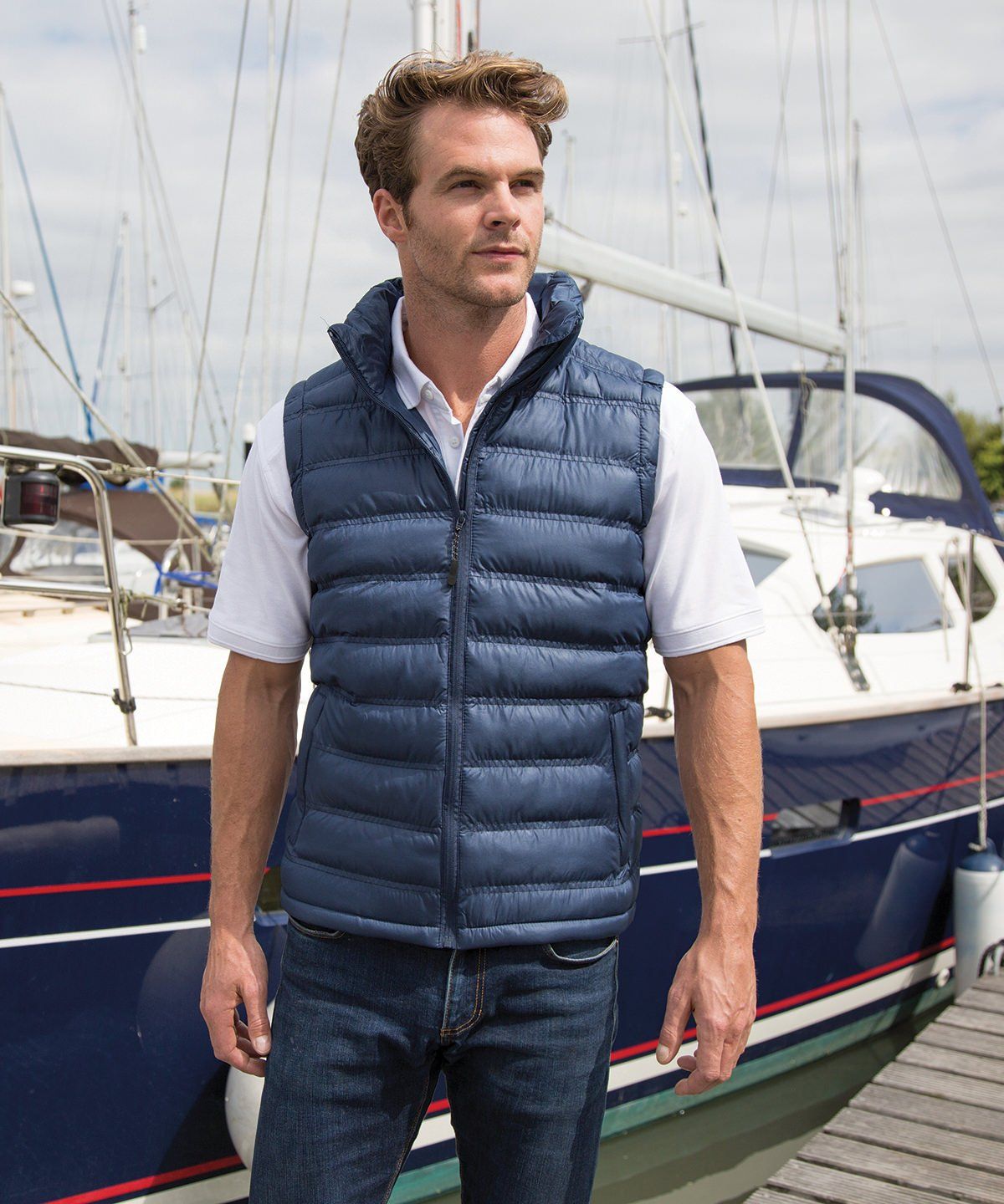WhiteLodge Man in navy puffer vest and jeans standing by a sailboat.