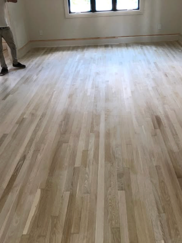 A person standing in an empty room with newly sanded, unfinished light-toned hardwood floors.