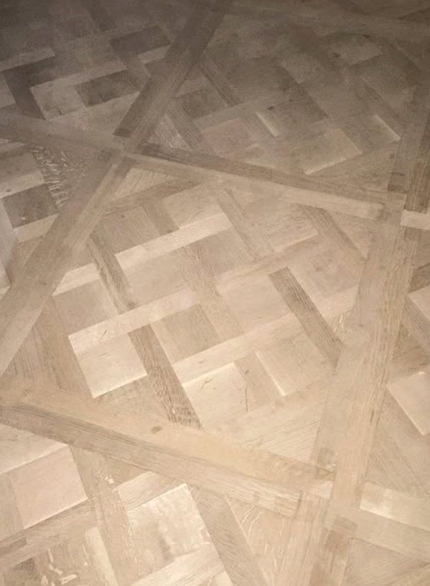 A light-colored wooden floor featuring a geometric pattern of interlocking rectangular panels and diagonal borders.