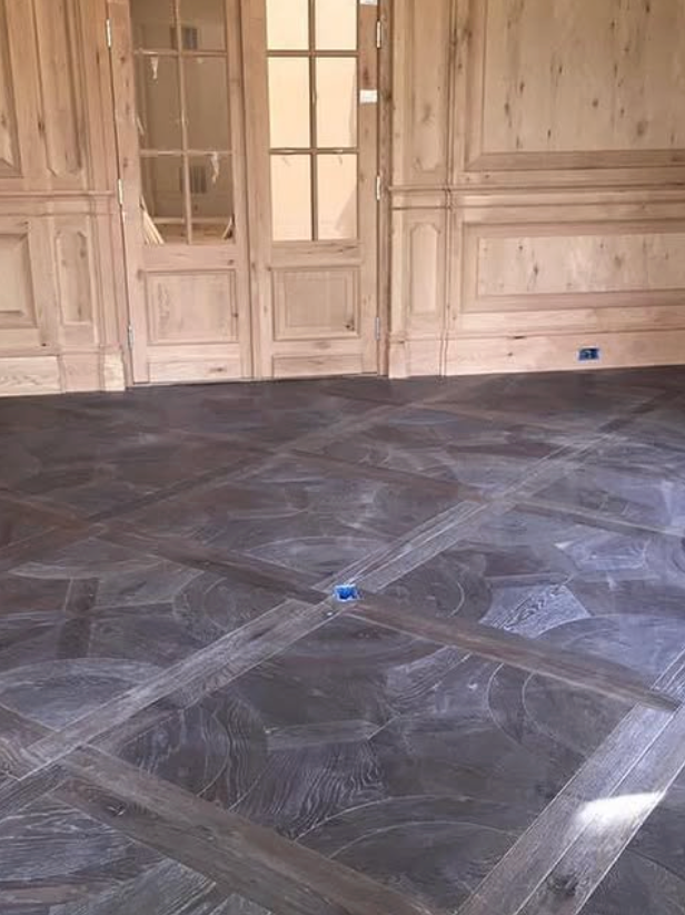 A room with light wood paneled walls and doors, featuring a geometric dark wood floor with visible sanding marks.