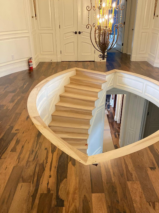 Heart-shaped stairwell opening in a wooden floor leading to the level below, in a room with white paneled walls.