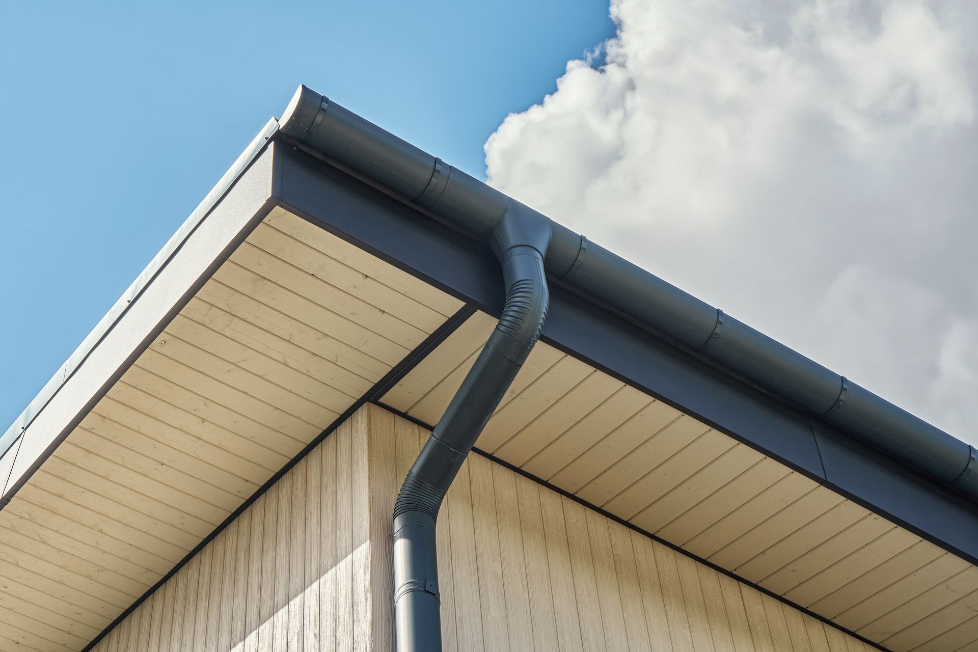 Corner of a building with dark gray gutters and downspout against a light blue sky with white clouds.
