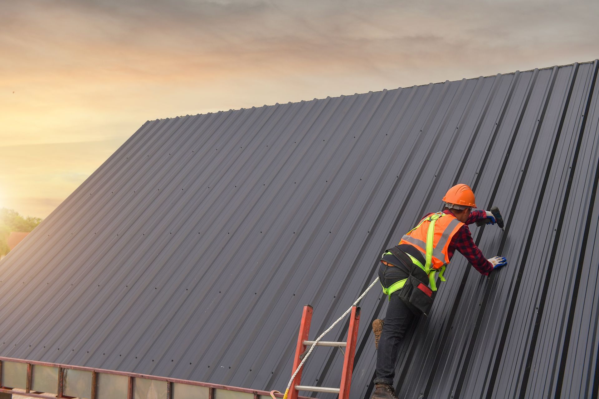 Roofer in orange vest and hard hat working on a dark gray metal roof, using a ladder in sunlight.