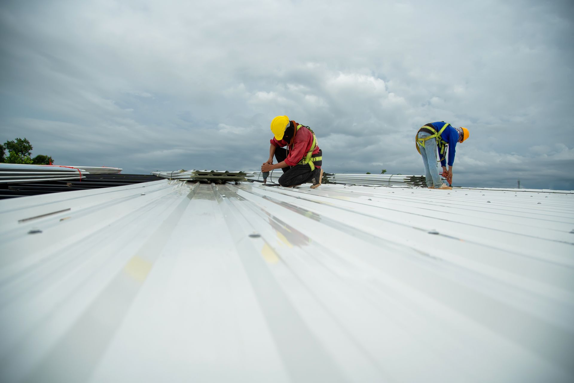 Two construction workers on a white metal roof, wearing hard hats and safety harnesses, working under a cloudy sky.