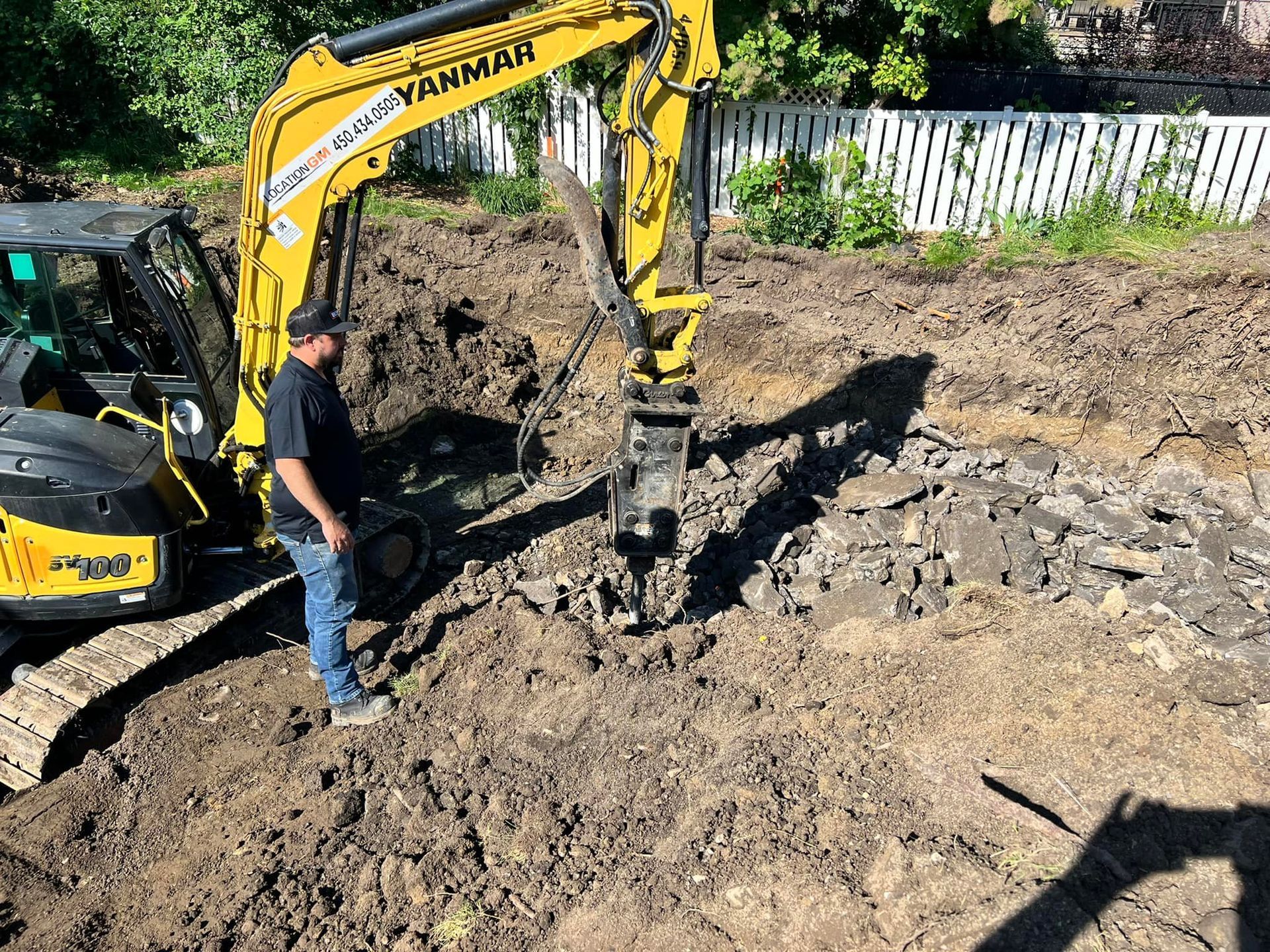 Une pelleteuse de chantier jaune est en activité près de l'entrée d'un bâtiment, des personnes se trouvant à proximité sous une tente et un cône de signalisation orange.