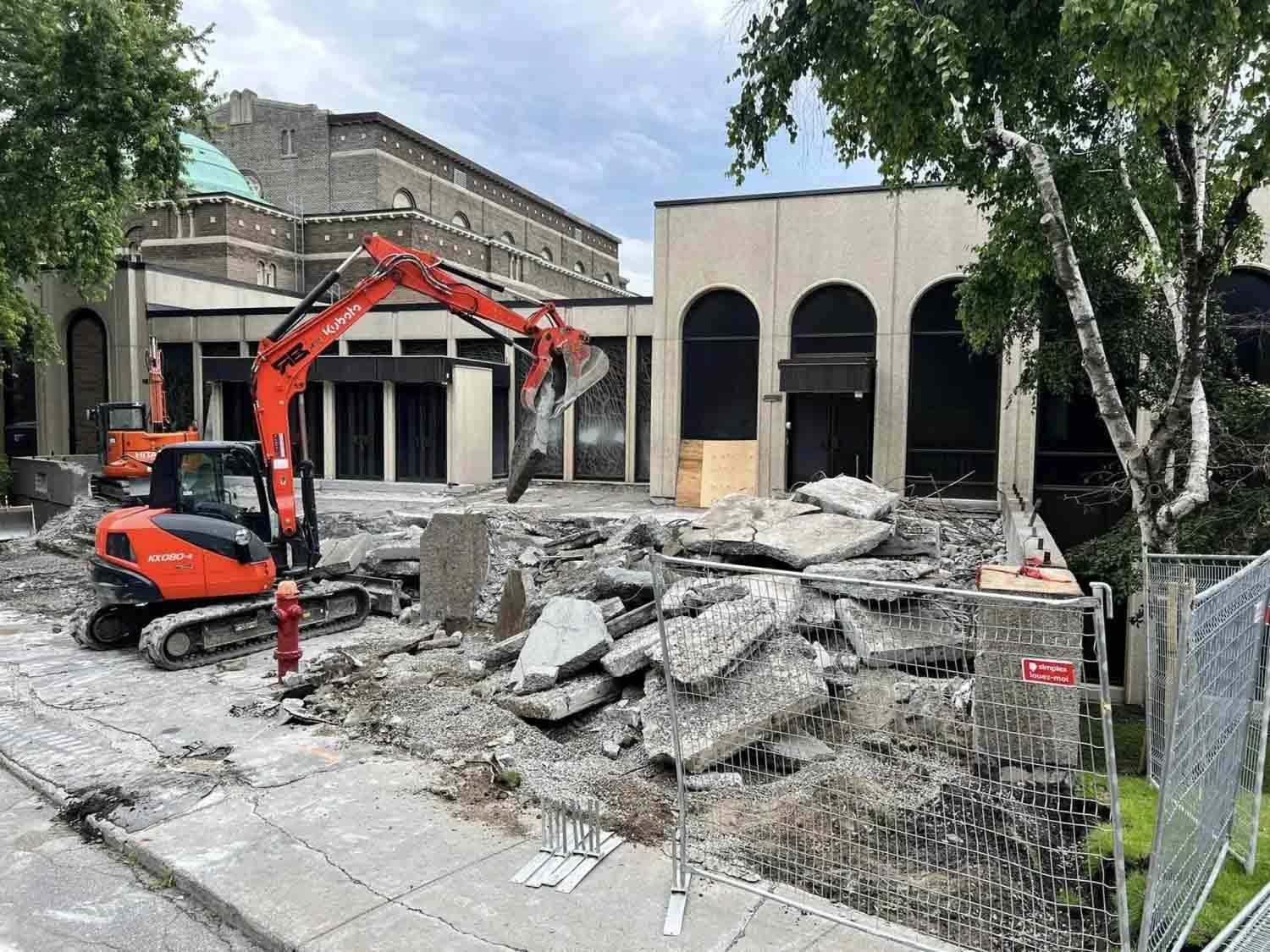 Une pelleteuse orange démolit un gros tas de gravats de béton devant un bâtiment aux fenêtres cintrées.