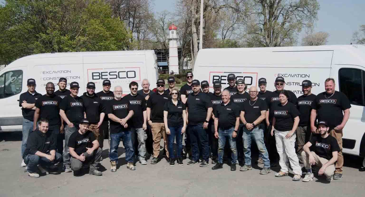 A group of workers in black Besco branded shirts pose for a photo in front of two white company vans on a sunny day.