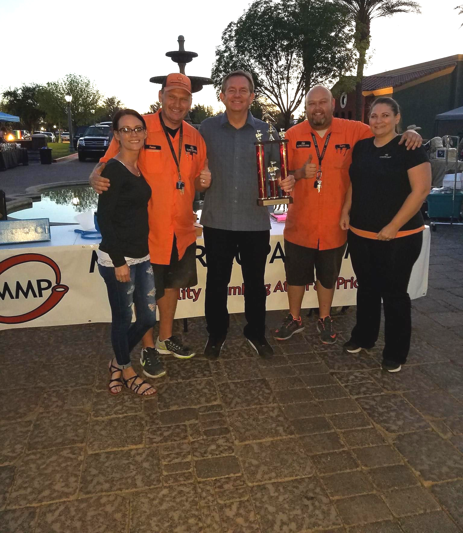 Group of five people, some in orange shirts, posing with trophy in front of a table outdoors.